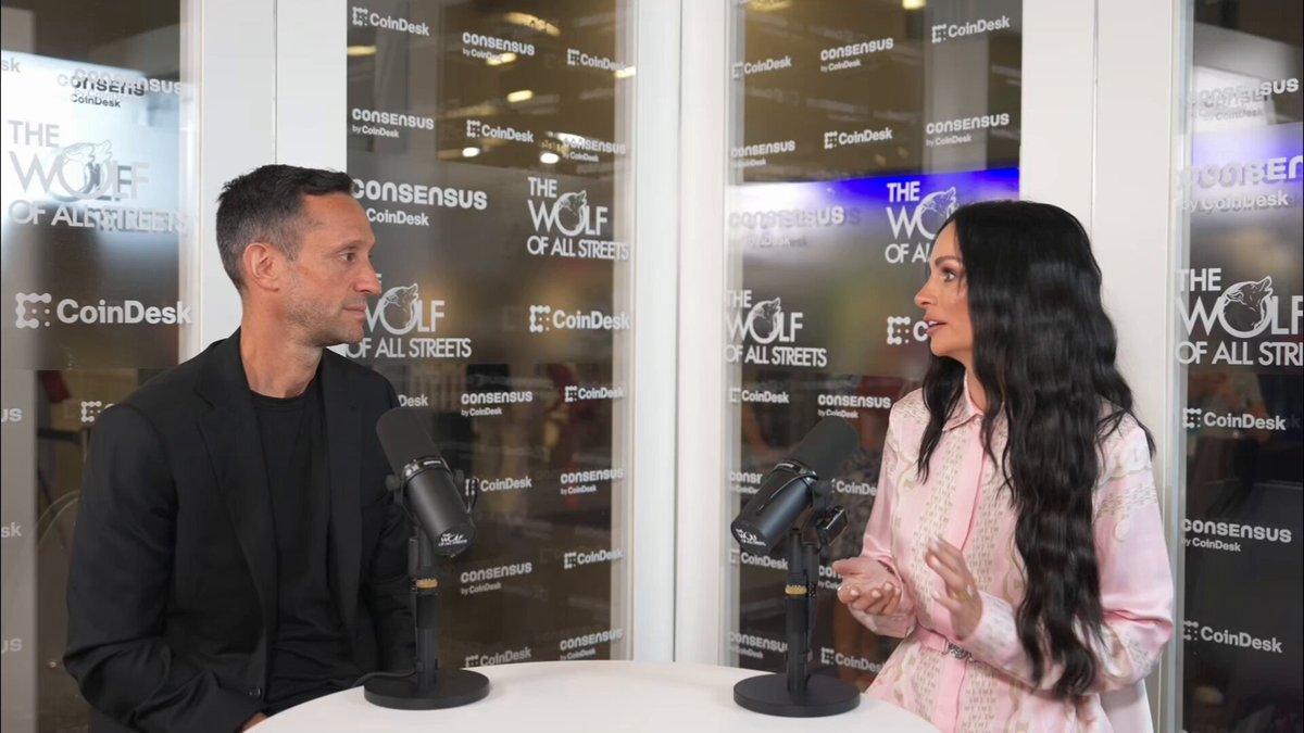 The image shows a man and woman sitting at a table, talking into microphones during an interview. The woman appears to be wearing a pink shirt while the man has dark hair. Both individuals are dressed professionally, with the man wearing a black suit jacket and the woman in a white blouse. They seem to be discussing something related to cryptocurrency or XRP, as indicated by the post they are talking about.