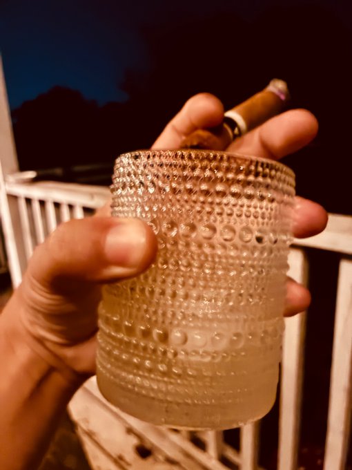First-person view of hands holding a textured glass with a clear beverage alongside a cigar, appearing to be on a balcony or porch at night.