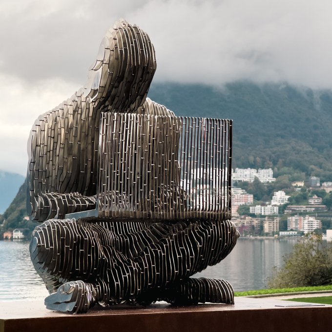 A large metallic sculpture of a hooded figure working on a laptop, mounted on a pedestal overlooking a lake with mountains and buildings in the misty background.