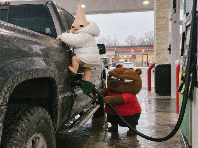 A humorous edited image showing a plush baby doll with a cone head hanging onto a muddy truck's side step while a plush brown bear character pumps gas into the vehicle.