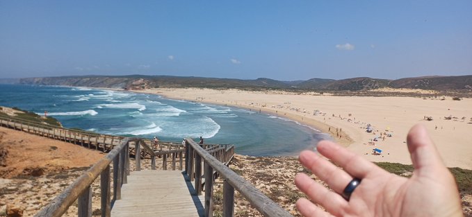 A scenic beach view from a wooden staircase leading down to the shore, with a person's hand and a black ring visible in the foreground.