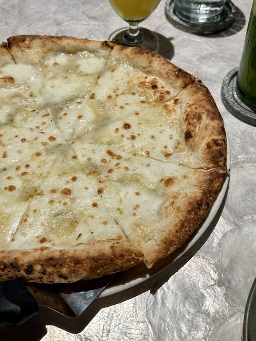 A close-up shot captures a freshly baked cheese pizza with a charred crust on a white plate, resting on a textured white tablecloth next to a glass.