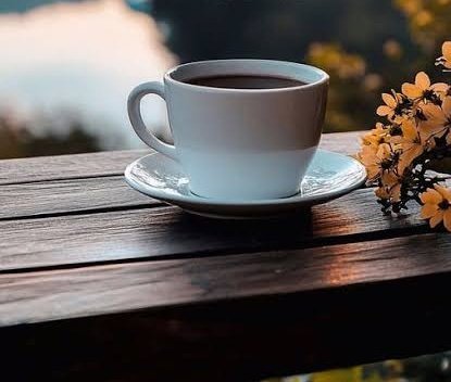 A white ceramic cup of black coffee on a saucer, placed on a wooden table with yellow flowers beside it, set against a blurred outdoor background.