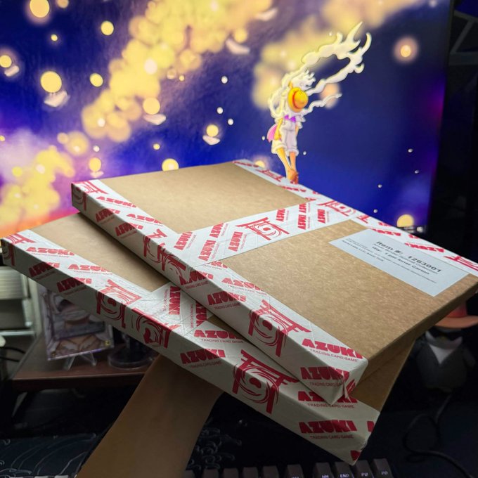 Two stacked cardboard boxes sealed with red and white Azuki "Trading Card Game" branded tape, sitting on a desk in front of a purple and yellow anime-style backdrop.