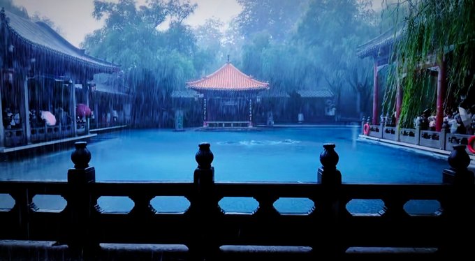 A nighttime view of a traditional Chinese-style hot spring pool illuminated in blue, featuring a pavilion with a red roof, rain falling, and a stone railing in the foreground.