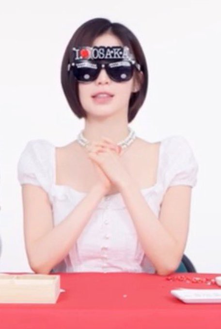 A woman with short dark hair wears a white puff-sleeve top, a pearl necklace, and sunglasses reading 'I ❤️ OSAKA', seated at a red table.