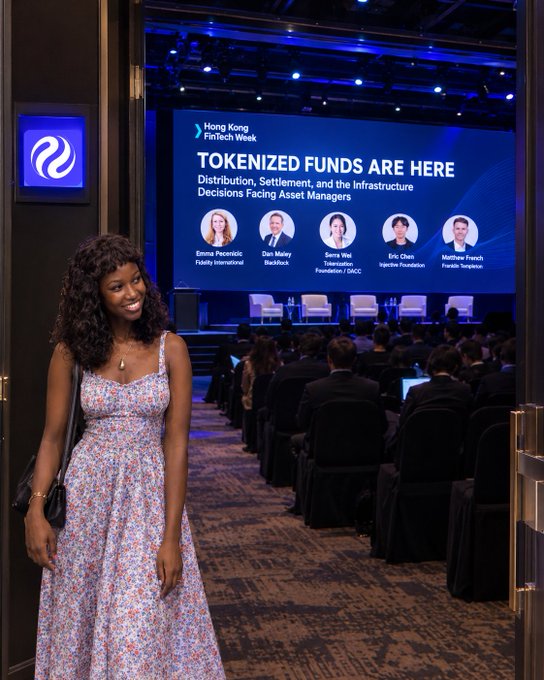 A woman in a floral dress poses in the doorway of a 'Hong Kong FinTech Week' conference, with a screen behind her titled 'TOKENIZED FUNDS ARE HERE' featuring speaker panels.