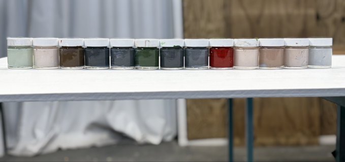 A row of small glass jars with white lids, filled with various colored powders and pigments, lined up on a white shelf.