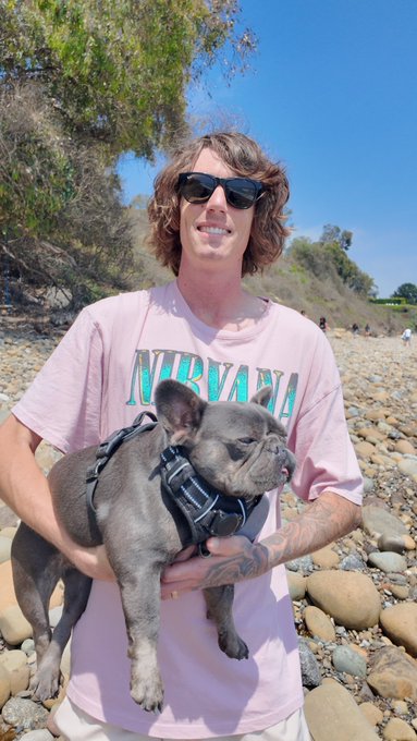 A smiling man with wavy brown hair wearing sunglasses and a pink Nirvana t-shirt, holding a gray French Bulldog with a plaid harness on a rocky beach.