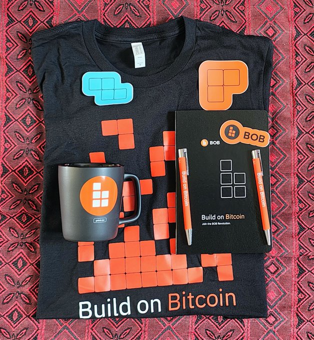 A flat-lay of BOB ("Build on Bitcoin") branded merchandise—including a black t-shirt with orange and teal tetromino designs, a gray coffee mug with the BOB logo, an orange sticker, and a notebook with "Build on Bitcoin / Join the BOB Revolution" text alongside two orange pens—rests on a patterned red textile, visually representing the expansion of the BOB community referenced in the "BOB OG army grows" post.