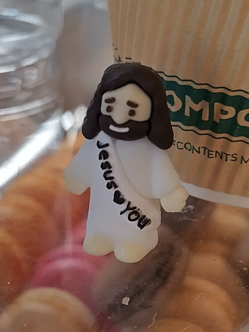 A small figurine of Jesus with 'Jesus loves you' written on its robe sits on top of what appears to be a pastry or food item in a container.