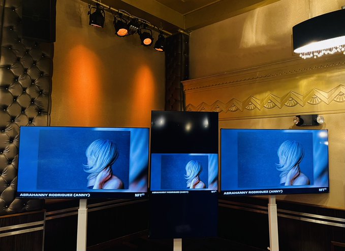 Multiple TV screens in an upscale venue displaying an anime-style avatar labeled 'ABRAHANNY RODRIGUEZ (ANNY)' with NFT branding.