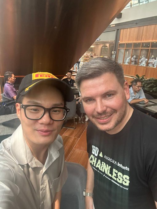 Two men pose for a selfie at a Token2049 event in Singapore, with one wearing a black "AURORA Intents - CHAINLESS" t-shirt connecting to the conference's crypto industry meetup described in the post.