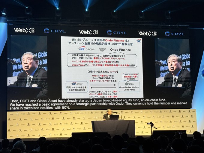 A conference stage with a presentation slide in Japanese about SBI Group and Ondo Finance. The visible English subtitle reads: 'Then, DGFT and Global Asset have already started a Japan broad-based equity fund, an on-chain fund. We have reached a basic agreement on a strategic partnership with Ondo. They currently hold the number one market share in tokenized equities, with 50%'.