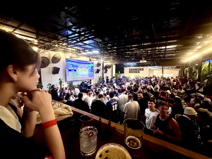 A crowded bar or venue with a large group of people watching a sports game on a projector screen, with drinks on a table in the foreground.
