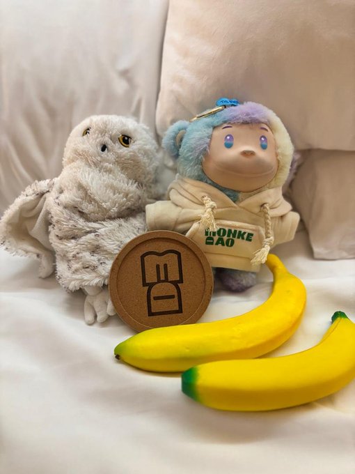 A small fluffy owl plush toy sits next to a colorful character plush wearing a hoodie that says 'THE MONKE BAG', with two yellow banana toys in front and a brown 'BD' circular object.