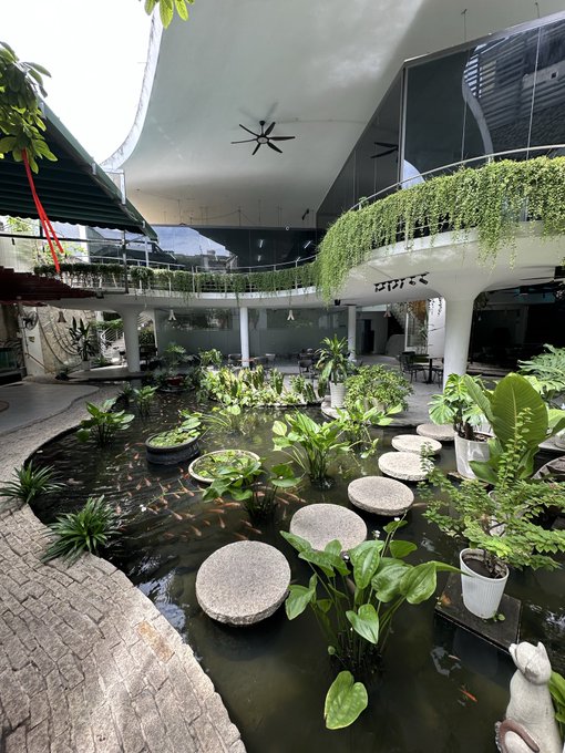 An indoor courtyard featuring a koi pond with lily pads, circular stepping stones, numerous potted plants, and a second-floor walkway adorned with hanging greenery, with a sign reading 'mực'.