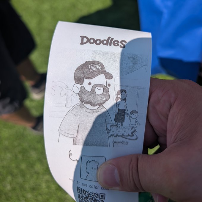A hand holding a "Doodles" collectible paper card featuring a black-and-white line drawing of a bearded character in a cap and a smaller figure in the background, with instructions to "scan to see color dood" and a QR code at the bottom.