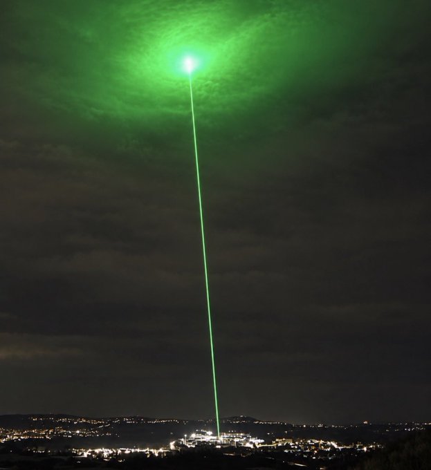 A bright green laser beam shoots from a brightly illuminated town into the night sky, lighting up the clouds above in a dramatic, otherworldly glow. The image visually mirrors the post's hype of an imminent, "huge" moment for memecoins, evoking a powerful, attention-grabbing signal breaking through the darkness of the crypto market.