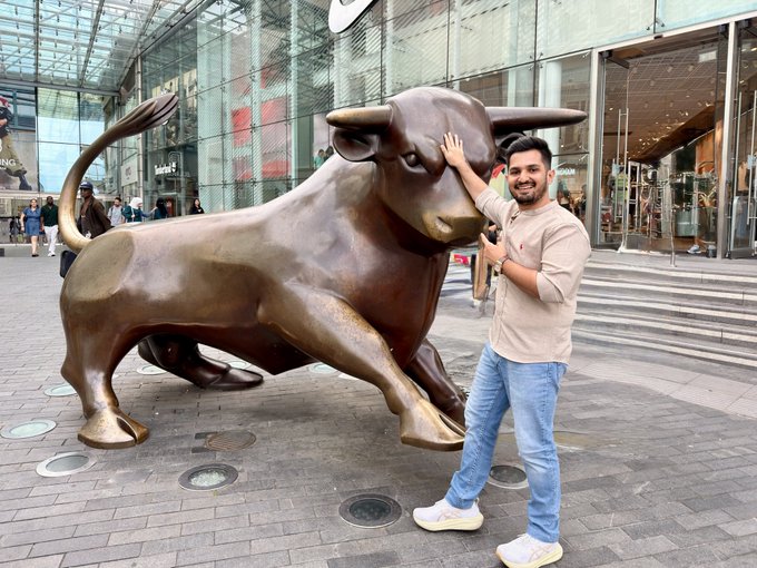A man smiles as he poses with his hand on the iconic bronze bull statue located outside the Frankfurt Stock Exchange (Börsenplatz). In the background, the "Bull and Bear" statues and a "Burger King" sign are visible, symbolizing financial markets and prosperity—fitting the caption's theme of being overwhelmed by a successful, prayed-for life.
