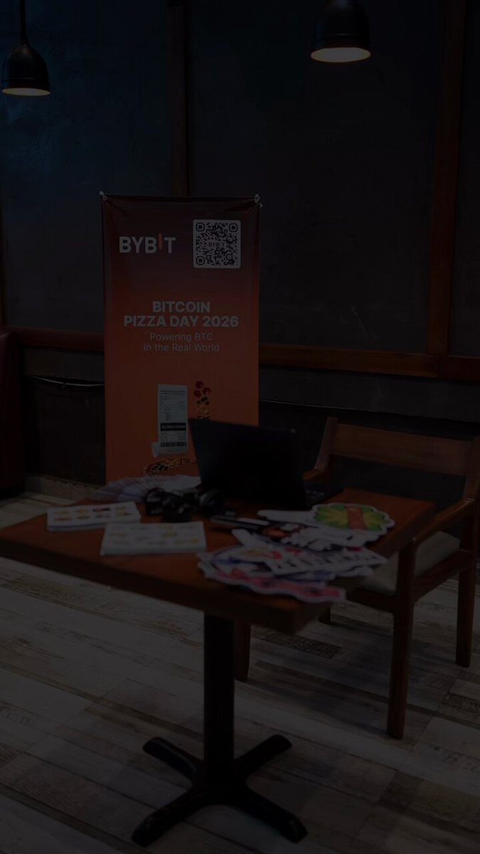 The image features a wooden dining table with various items placed on top of it. There are two chairs positioned around the table - one near the left side, and another towards the right side. On the table, there's an open laptop computer, along with some papers or documents. A pizza box is also present on the table. The background shows a dark room with a sign that reads "BYBT Pizza Day 2008".