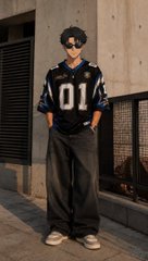 A young man posing on a street wearing sunglasses, a black and white '01' jersey, loose dark pants, and sneakers.