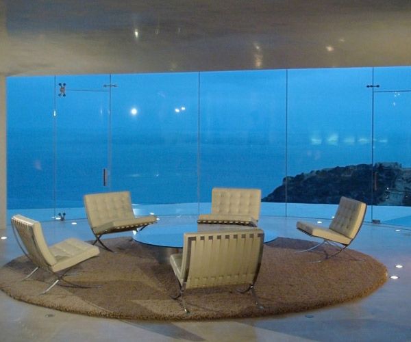 The image features a modern, circular room with large windows overlooking an ocean view. The room contains six white chairs arranged around a round coffee table, creating a comfortable seating area for conversation or relaxation. The chairs are positioned at various angles, giving the impression of a dynamic and inviting space.