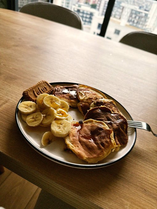 A plate of pancakes with chocolate spread and sliced bananas, along with a Lotus Biscoff cookie, served on a wooden table near a window overlooking a city.