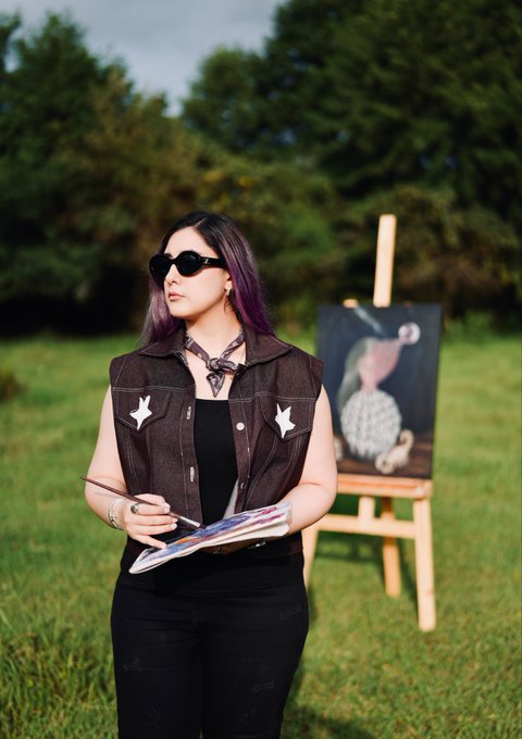 A woman with purple-tinted hair and sunglasses stands outdoors holding a palette and brush, with a painting displayed on an easel behind her.