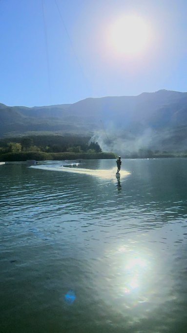 A person waterskiing across a calm lake with mountains and bright sun in the background, leaving a trail of mist behind them.