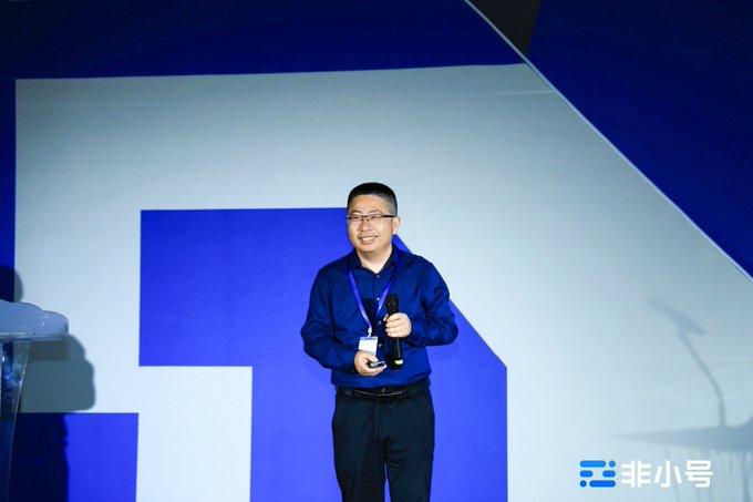 A man in a blue shirt with a lanyard speaking on stage while holding a microphone, with a blue and white geometric backdrop and a watermark reading '非小号' in the corner.