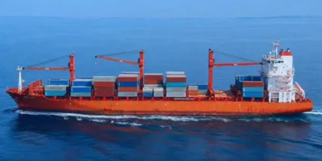 A red and white cargo ship loaded with colorful shipping containers sailing on calm blue ocean waters, featuring tall cranes and a multi-story bridge structure at the stern.