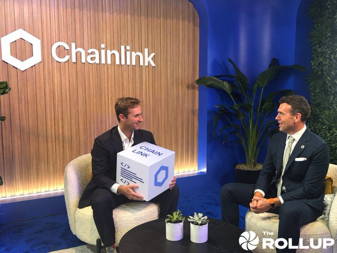 Two men in suits sit in chairs on a stage with a Chainlink logo on the wall behind them; one holds a Chainlink cube. The Rollup logo appears in the corner.