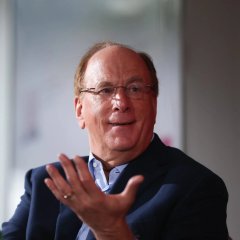 A bespectacled man in a blue blazer speaks while gesturing with his hand, likely captured during a conference or panel discussion. The image accompanies the post about BlackRock's expanding role in the crypto space, visually representing a key figure from the financial industry shaping the emerging digital asset landscape.
