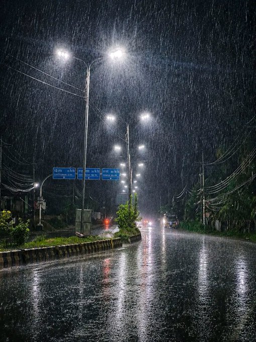 A nighttime photograph of a wet, rain-slicked road illuminated by tall streetlights, with rain visibly falling and reflections shimmering on the pavement.