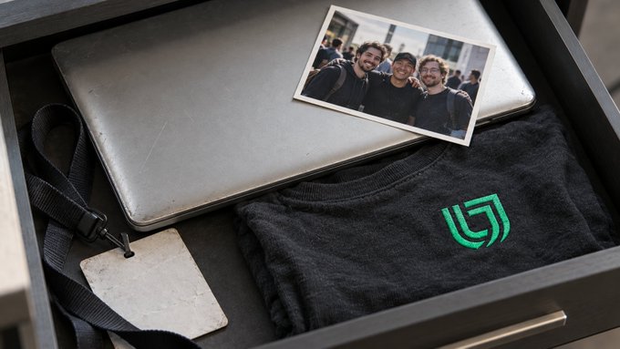 An open drawer contains a black laptop, a photo of three smiling men, a black t-shirt with a green logo, and a blank name tag attached to a lanyard.