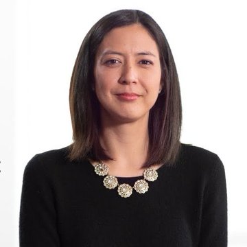 A woman with shoulder-length brown hair wearing a black top and an ornate crystal flower necklace poses for a portrait against a white background.