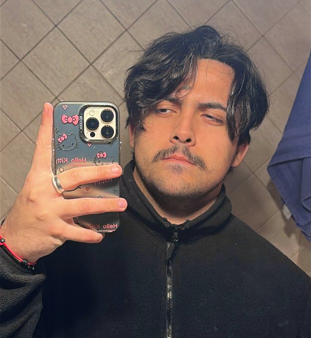 A young man with tousled dark hair and a light beard takes a mirror selfie in a tiled bathroom, wearing a black zip-up hoodie and holding a phone with a "Hello Kitty" case featuring pink bows. His relaxed expression and casual pose pair with the post's "gm beloved followers" greeting, making it a friendly morning selfie for his audience.
