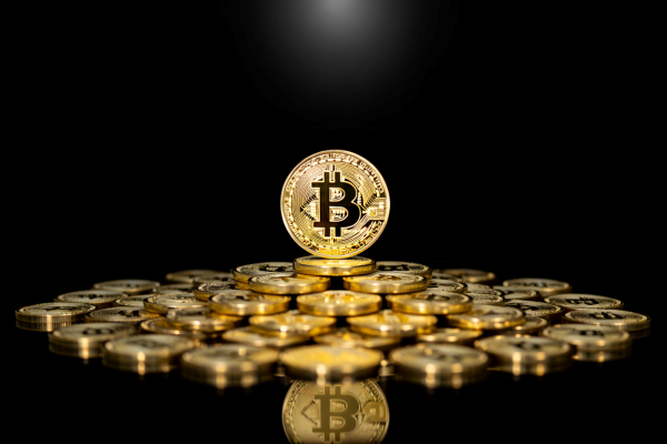 A stack of golden Bitcoin coins arranged in a pyramid formation with one coin standing upright at the center, prominently displaying the "₿" symbol against a black background, visually representing the post's message about Bitcoin's milestone of having 20.07 million mined coins—95.6% of its total supply—and its role as a deflationary asset in the era of stablecoins and blockchain trading.