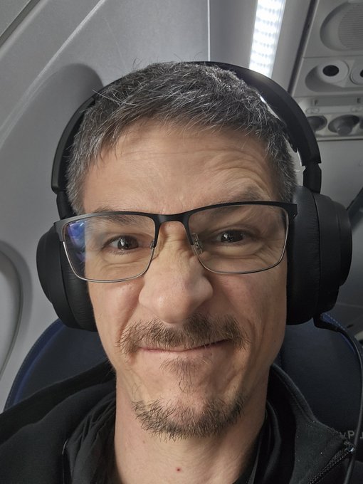 A close-up selfie of a man with short grey hair, glasses, and a goatee wearing large black over-ear headphones inside an airplane cabin.
