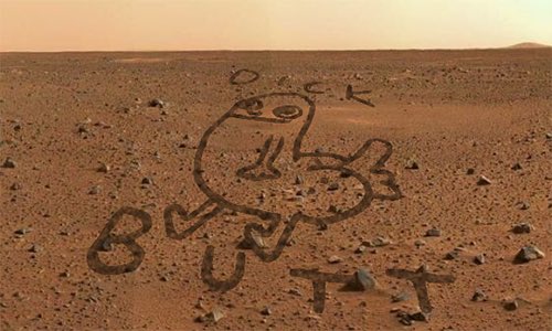 An overhead view of a Mars-like rocky landscape where pebbles are arranged to spell 'BUTT' alongside a drawing of a cartoonish figure.
