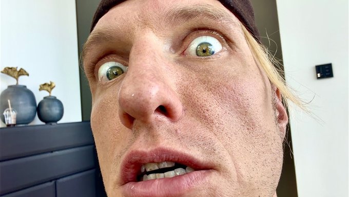 A man with a shocked, wide-eyed expression looks directly into the camera in a close-up selfie, with decorative vases visible on a shelf behind him. The image's exaggerated facial expression visually conveys the urgent tone suggested by the post's call for "URGENT" new Bitcoin trades.