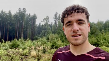 A man wearing a maroon athletic shirt stands outdoors in front of a forest of pine trees.