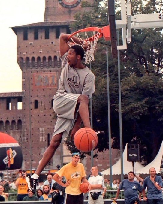 A basketball player in a grey Adidas outfit performs a slam dunk on an outdoor court, with a historic brick tower and spectators visible in the background.
