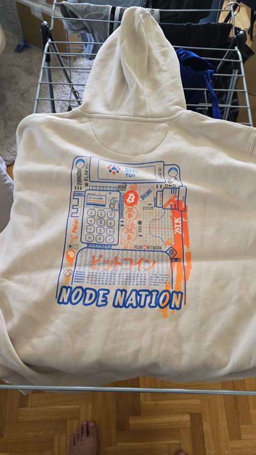The image features a white hoodie with blue and orange designs, including a computer screen and a calculator. The hoodie has the text "NODE NATION" written on it. The hoodie appears to be hanging from a clothes dryer or drying rack in front of a wooden floor.