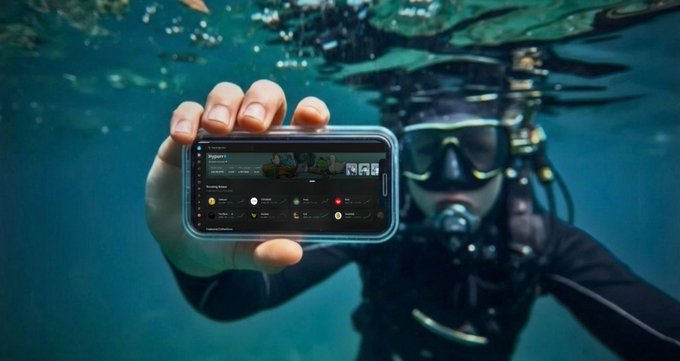 An underwater photograph of a scuba diver holding a waterproof phone case displaying a chat or social media interface toward the camera.