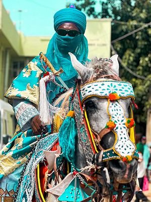 A person wearing a teal turban and face covering rides a horse elaborately decorated in matching teal and gold ornamental regalia.