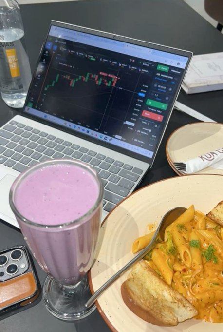 A breakfast setup featuring a laptop displaying stock/crypto trading charts alongside a purple smoothie and pasta dish, captioning "Gm" as a casual morning greeting to fellow traders starting their day with market analysis over brunch.