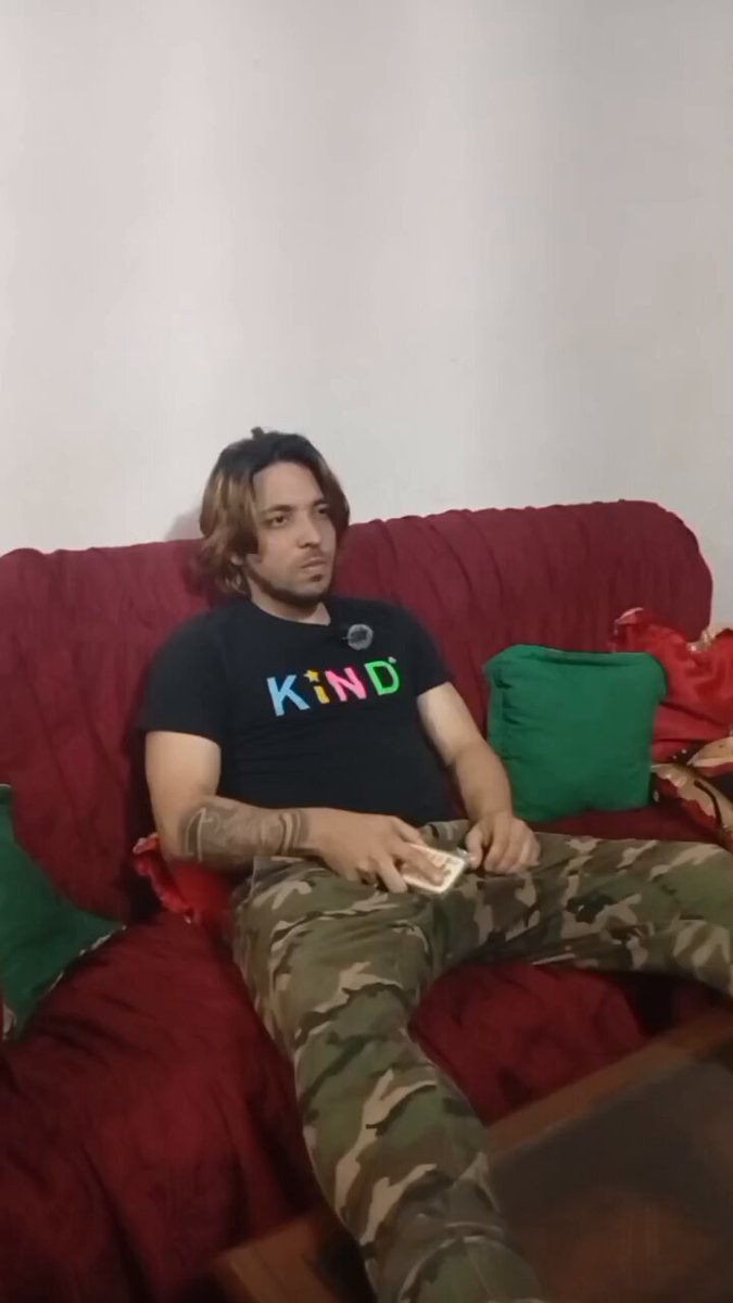 The image features a man sitting on a red couch with his legs crossed, wearing a black shirt that says "kind" on it. He has long hair and is holding a cell phone in his hand. The background of the photo shows another person's face, but only a small portion of their upper body can be seen.