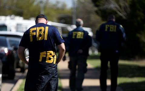 Several FBI agents wearing dark blue jackets with large yellow 'FBI' lettering on the back walk away from the camera down a sidewalk.
